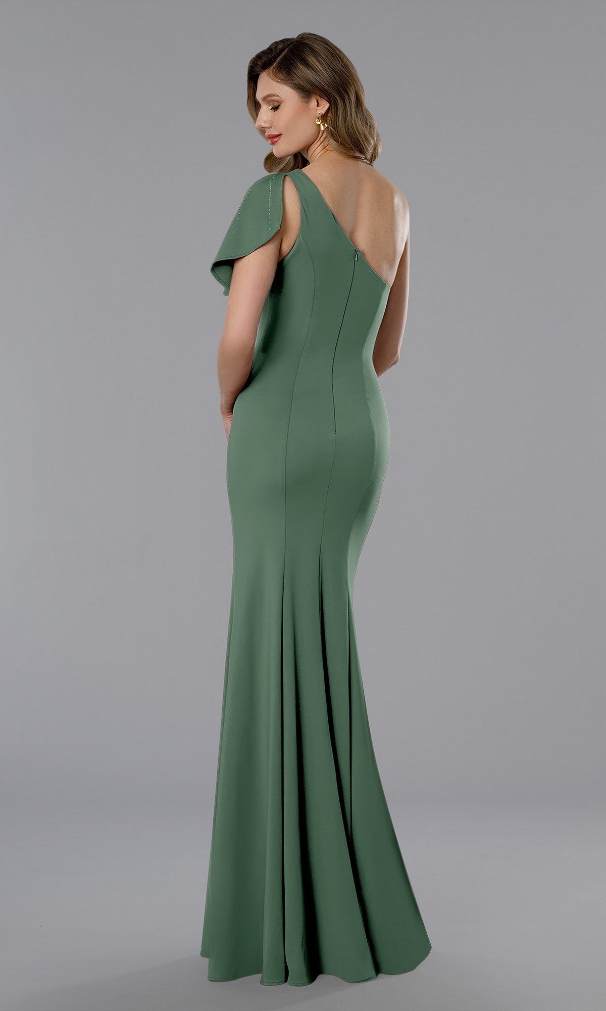 A woman in a Stella Couture 25358 long formal dress in sage, featuring a fitted, floor-length silhouette, one-shoulder asymmetrical neckline, and back zipper closure stands against a plain gray background.